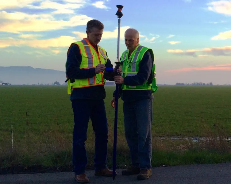 Two surveyors checking a GNSS rover in a field at sunset